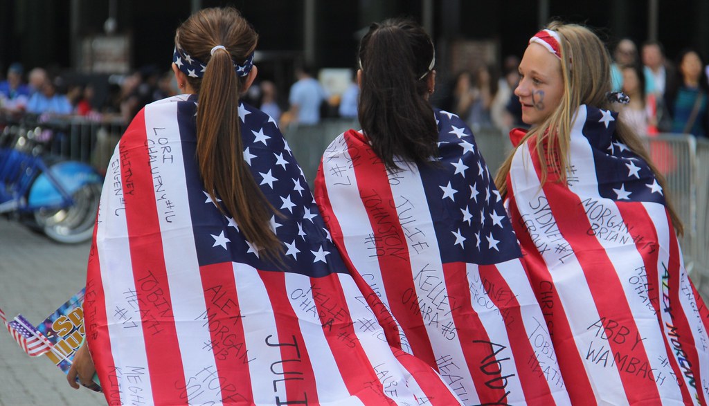 A group of young soccer fans with the American flag draped over them.