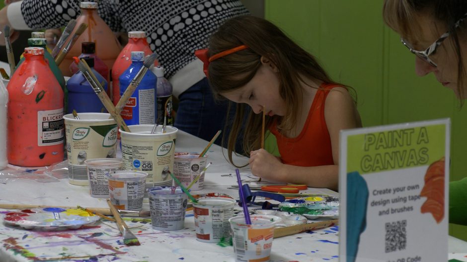 A little girl in a red dress sits at a table covered in painting supplies, painting something on a small canvas.