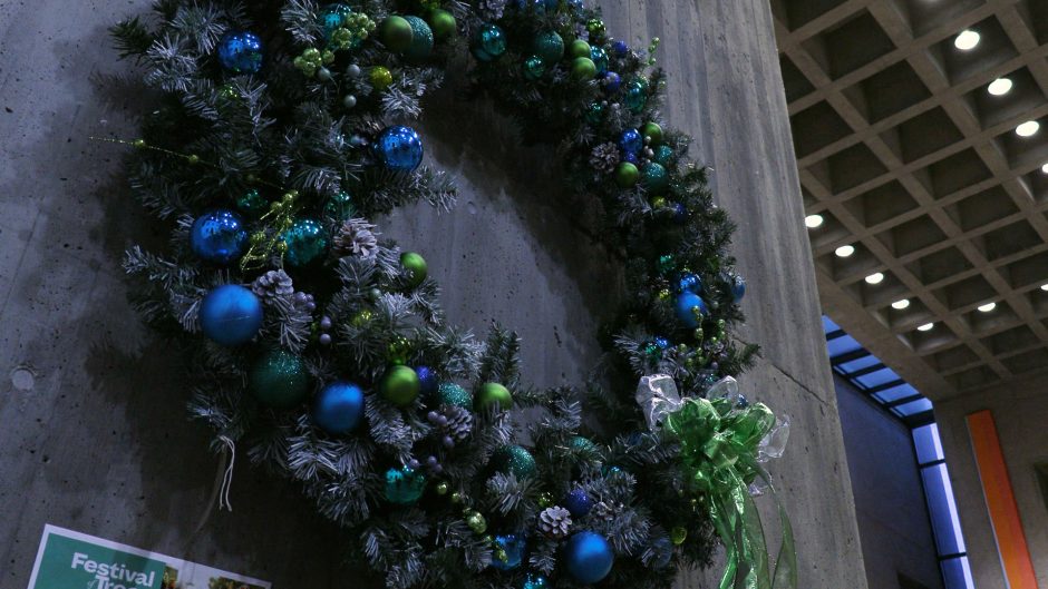 A giant blue, green, and silvery Christmas wreath hangs on a gray concrete wall.
