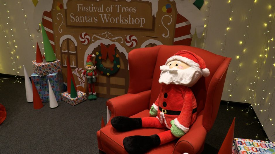 A large Santa Claus plushie sits in a cozy red chair in front of a backdrop that reads "Festival of Trees: Santa's Workshop".
