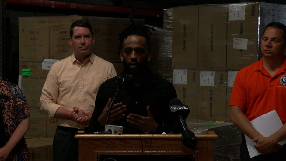 A man stands at a podium with three people behind him and cardboard boxes in the background.