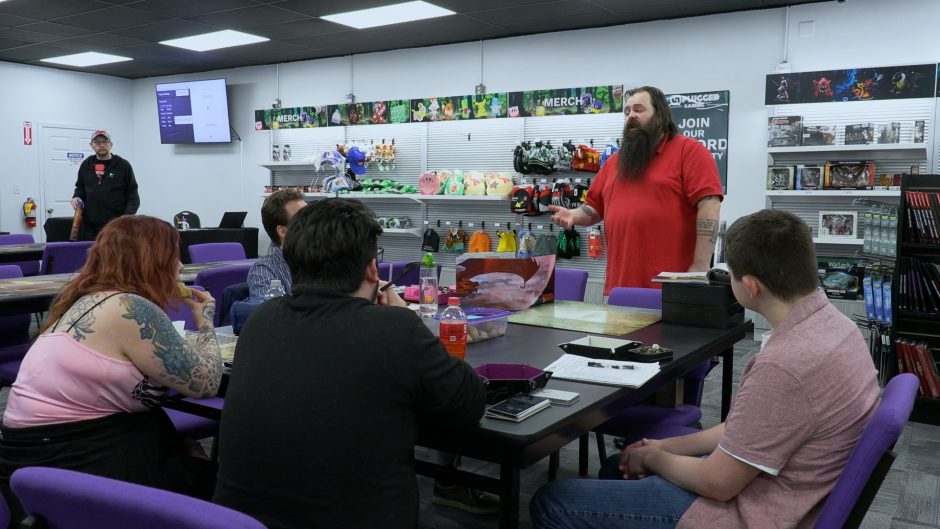 A group of people sit around a table with a man in a red shirt and large beard addressing the group.