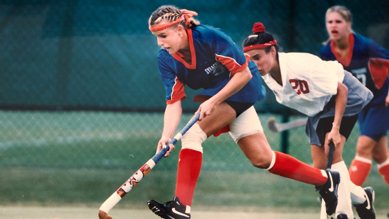 Women in blue shirt and orange socks, reaches out to play the ball with her stick