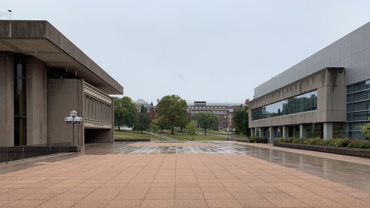 Dark rainclouds are above Syracuse University's Newhouse building. The rain is coming down and there are puddles already forming on the ground.