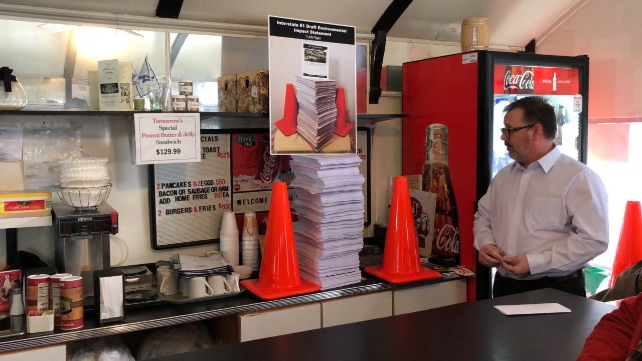 A man in a white shirt stares at a multiple-feet-tall stack of papers flanked by two orange traffic cones. The papers and the cones are behind the counter of a diner.