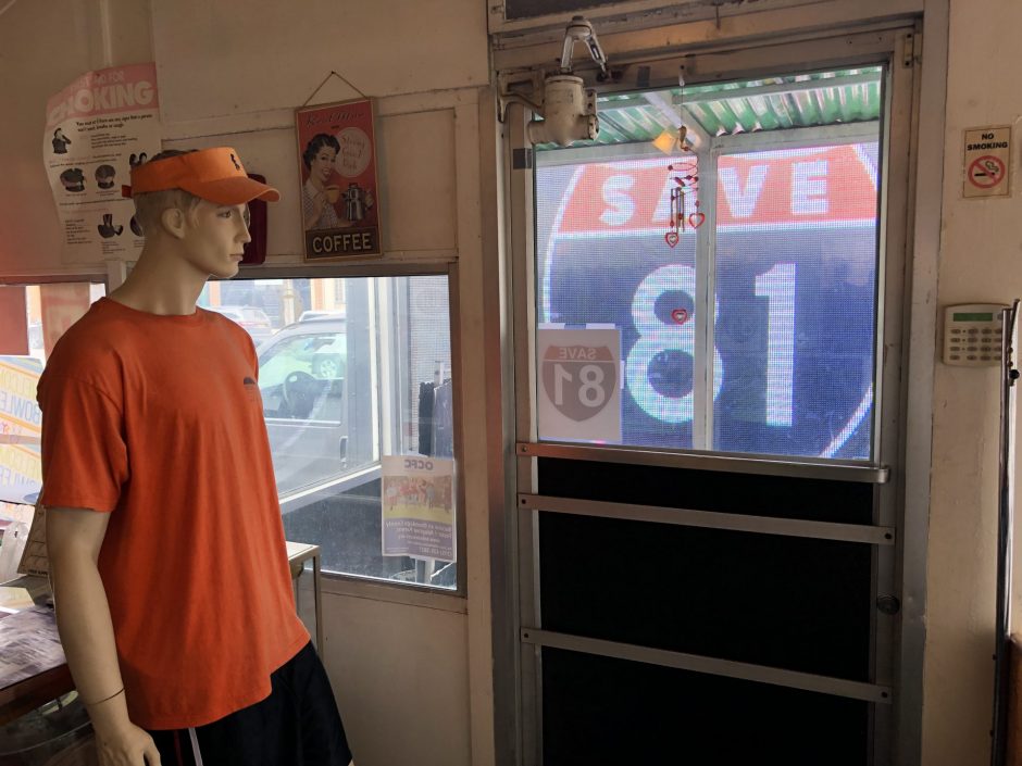 A male mannequin wearing an orange shirt and visor stares at the front door of a diner. The windows are filled with a large, illuminated sign that reads "Save 81".