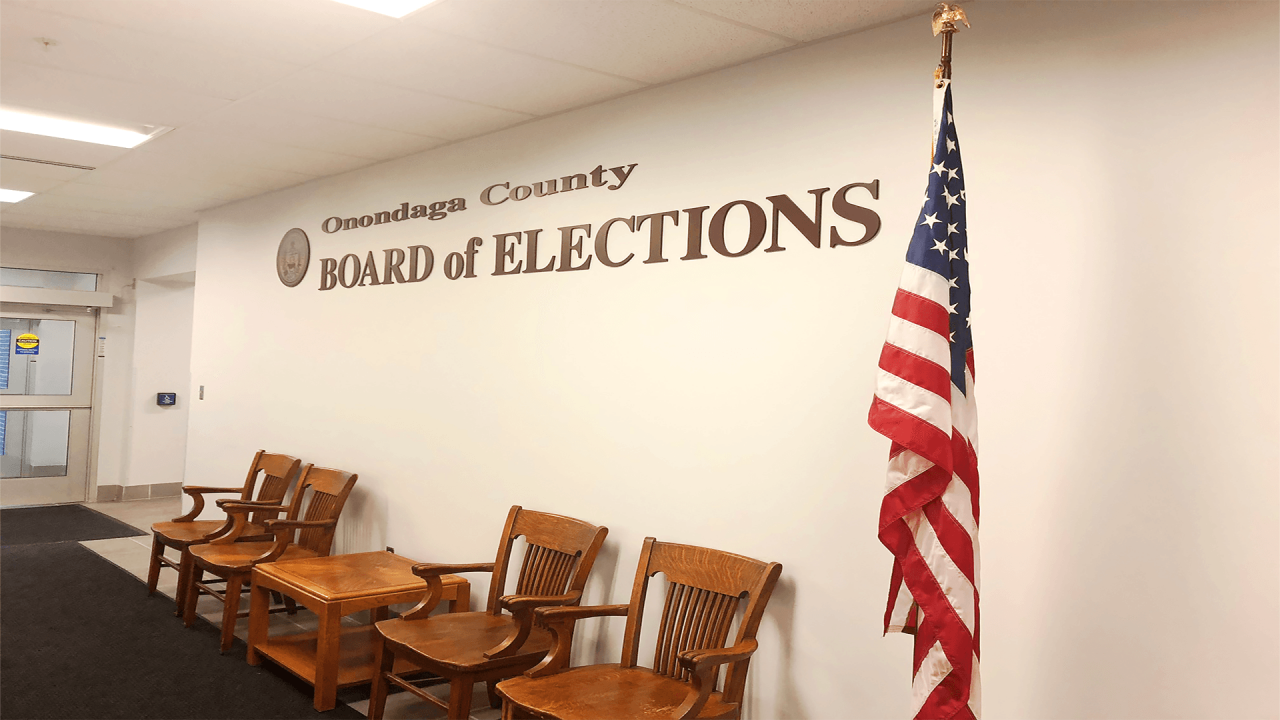Wall with "Onondaga County Board of Elections". A few chairs in front and an American flag.