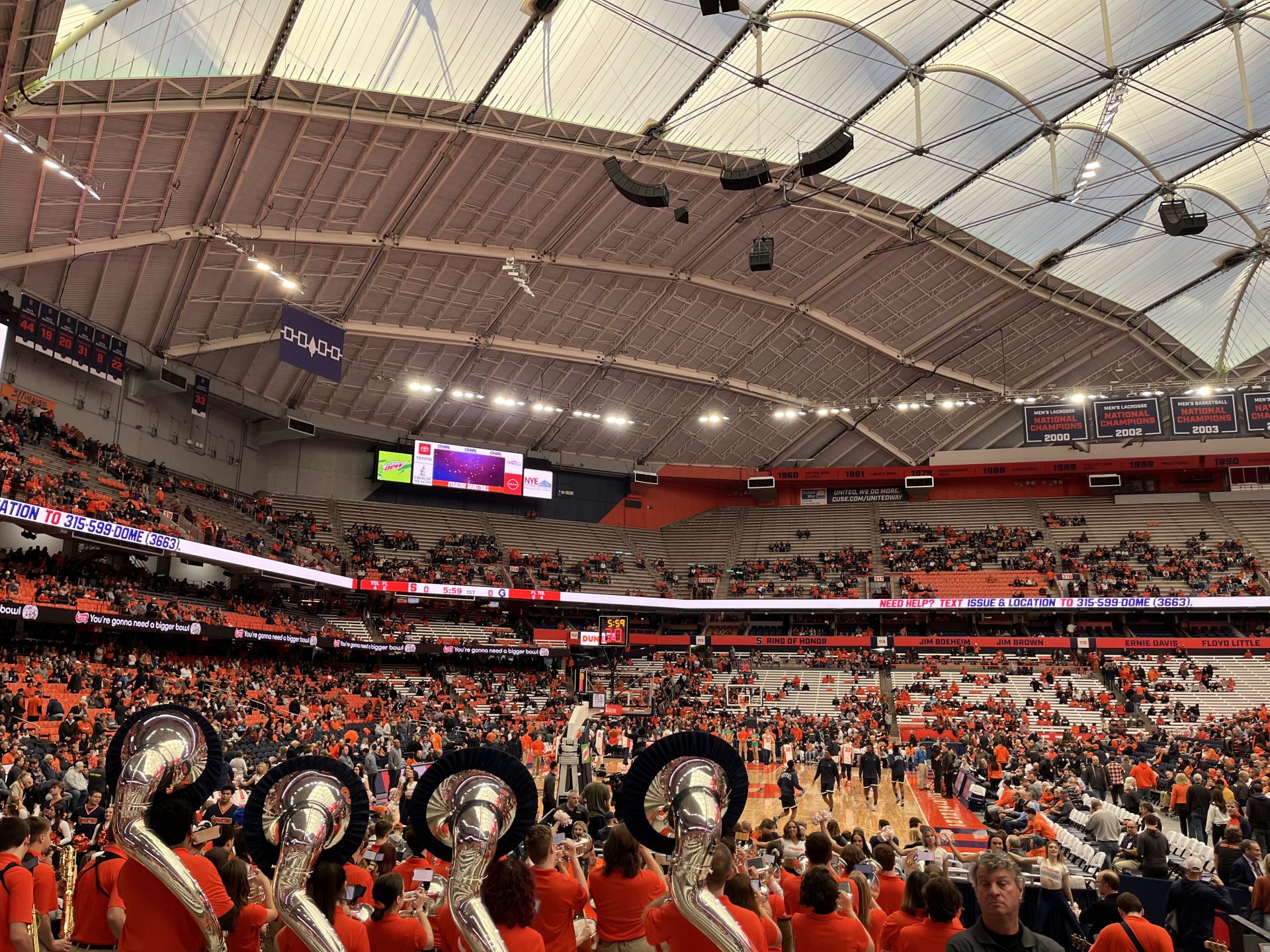 A crowd of people dressed in Syracuse Orange surround a tan basketball court with a grey dome above and a music band at the bottom of the image.
