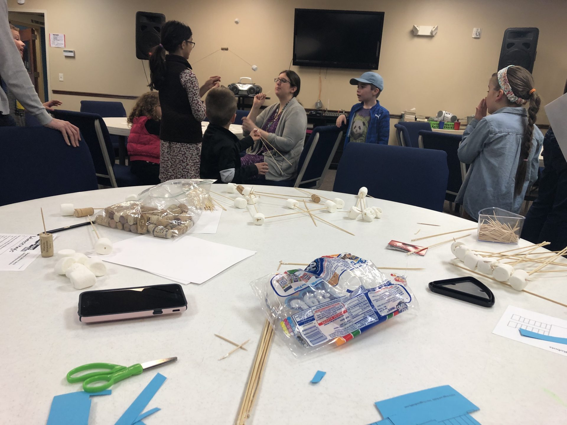 Table with paper, marshmallows, wood skewers and paper laid across. Students, parents and teacher are standing and sitting in the background.
