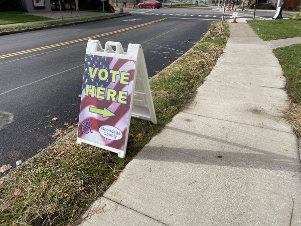 A sign sitting on a curb that says "Vote Here", with an arrow pointing toward a polling place.