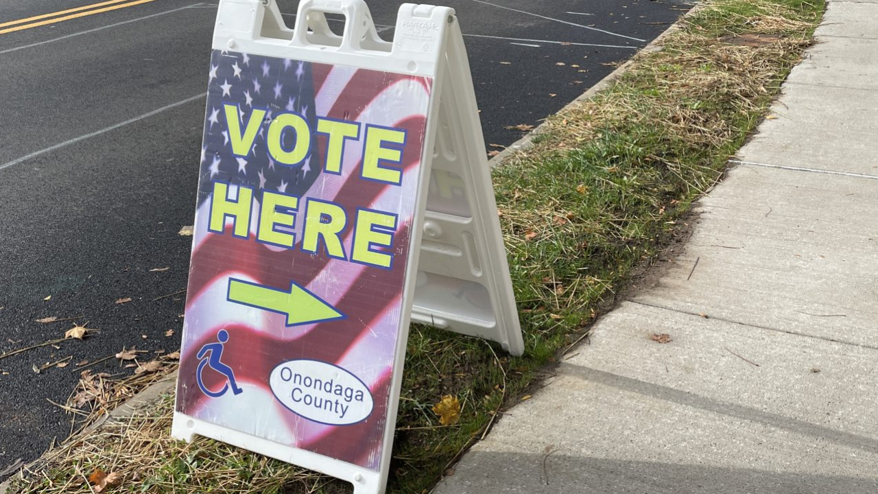 A "Vote Here" sign sits on a curb, with an arrow pointing to a public library, to indicate where citizens can vote early.