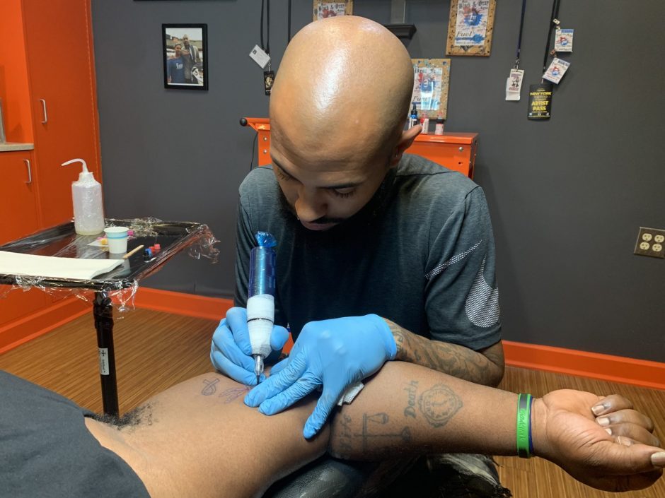 Dashaun Smith wears blue surgical gloves and is holding a blue tattoo machine. He is tattooing a dragonfly on another mans inner arm.