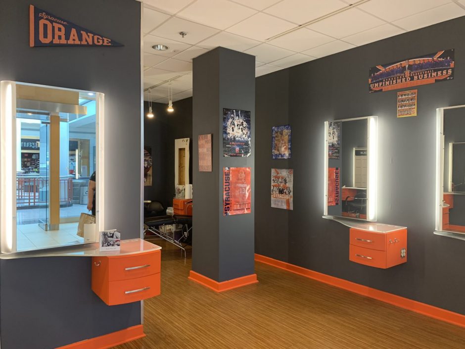 One area of the 'Cuse Ink shop. It is painted dark gray with orange trim. There are three mirrors that are backlit. Various Syracuse University banners and posters hang around the shop.