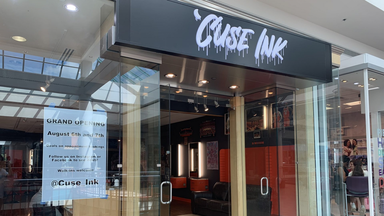 The mall storefront of 'Cuse Ink. The doors and windows are clear glass. In the left window there is a sign that reads "GRAND OPENING. August 6th and 7th. Deals on appointment bookings. Follow us on Instagram or Facebook to see details! Walk-ins welcome. @Cuse_Ink." Above the store is a sign with a black background and white light-up text in a dripping font that reads "'Cuse Ink."