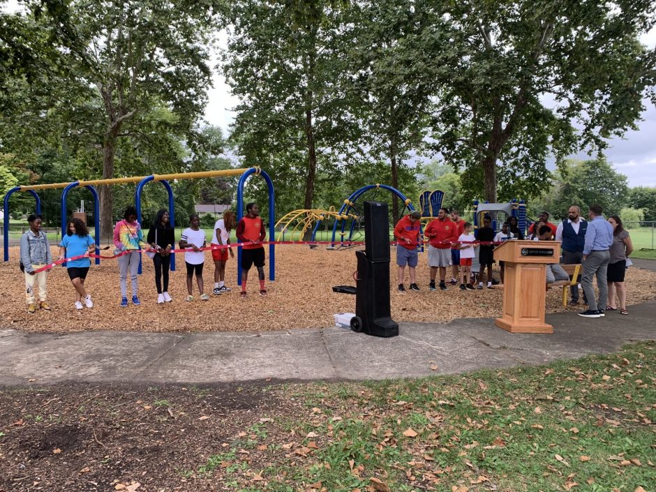 A line of young people stand holding a ribbon. Behind them are trees and a playground. In front of them is a speaker and a podium.