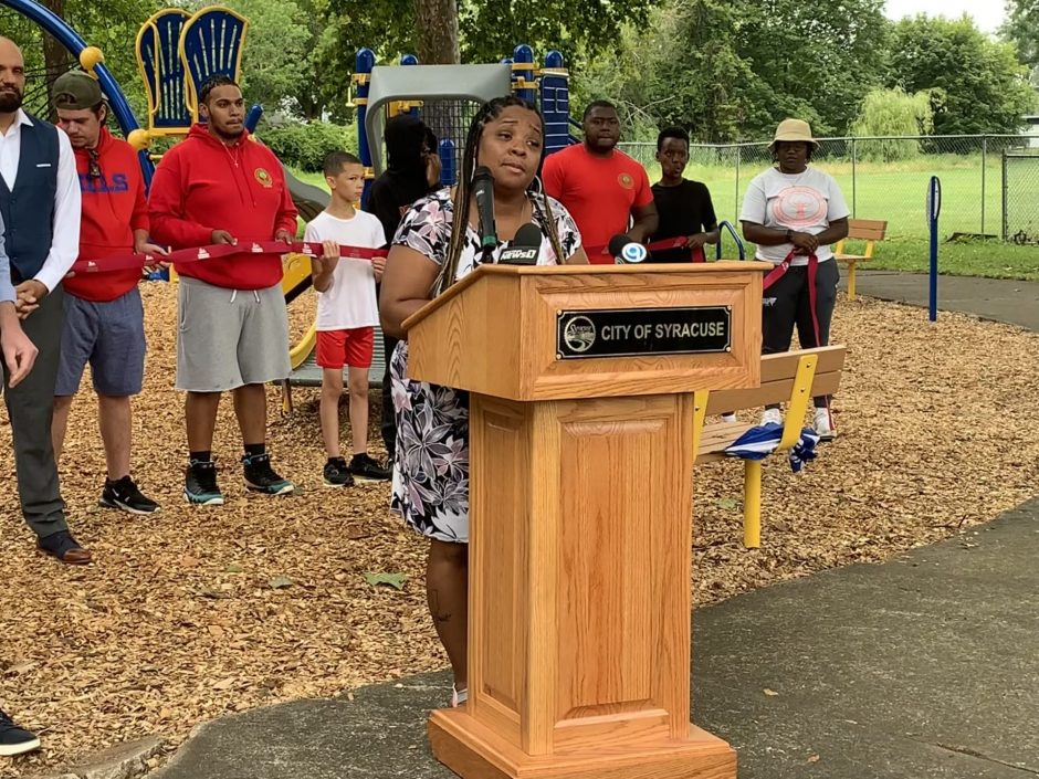 Rasheada Caldwell stands behind a podium speaking into a microphone. Behind her is a line of people holding up a red ribbon.