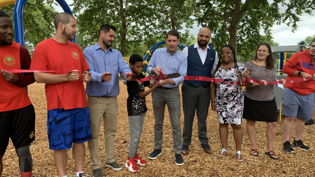 A group of people stand in a line holding a red ribbon. It the middle of the line, a child and Mayor Ben Walsh hold a large pair of scissors and cut the ribbon. In the background are trees and the park.
