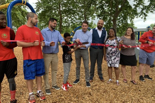 A group of people stand in a line holding a red ribbon. It the middle of the line, a child and Mayor Ben Walsh hold a large pair of scissors and cut the ribbon. In the background are trees and the park.