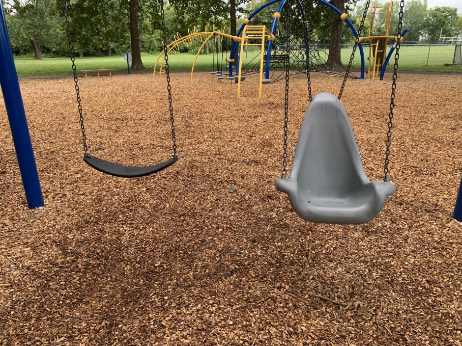 Two swings hang from a swing set. The swing on the left is a traditional swing. The swing on the right is a handicap accessible swing.