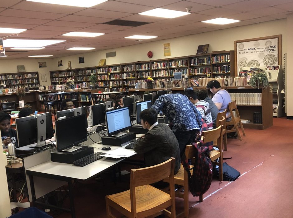 High School students in the library with bookshelves against the walls typing at a computer cluster.