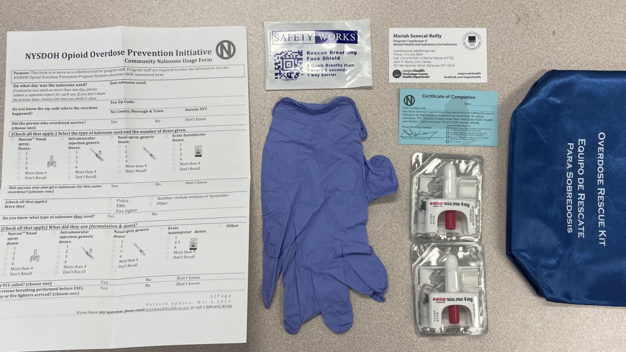 This photo shows the contents of a Narcan kit. Inside a pouch are two Narcan nasal sprays, a Rescue Breathing Face Shield, a pair of rubber gloves, and instructions on how to give Narcan to someone.