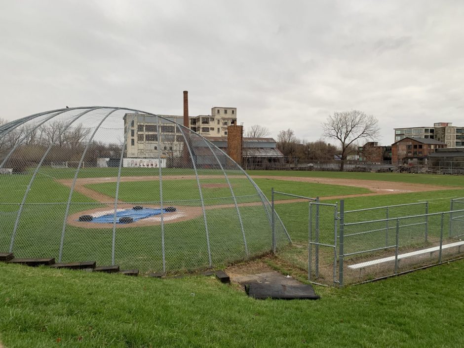 A wide view of the entire Fowler High School baseball field from behind home plate, showing a crumbling factory beyond the center field fence.