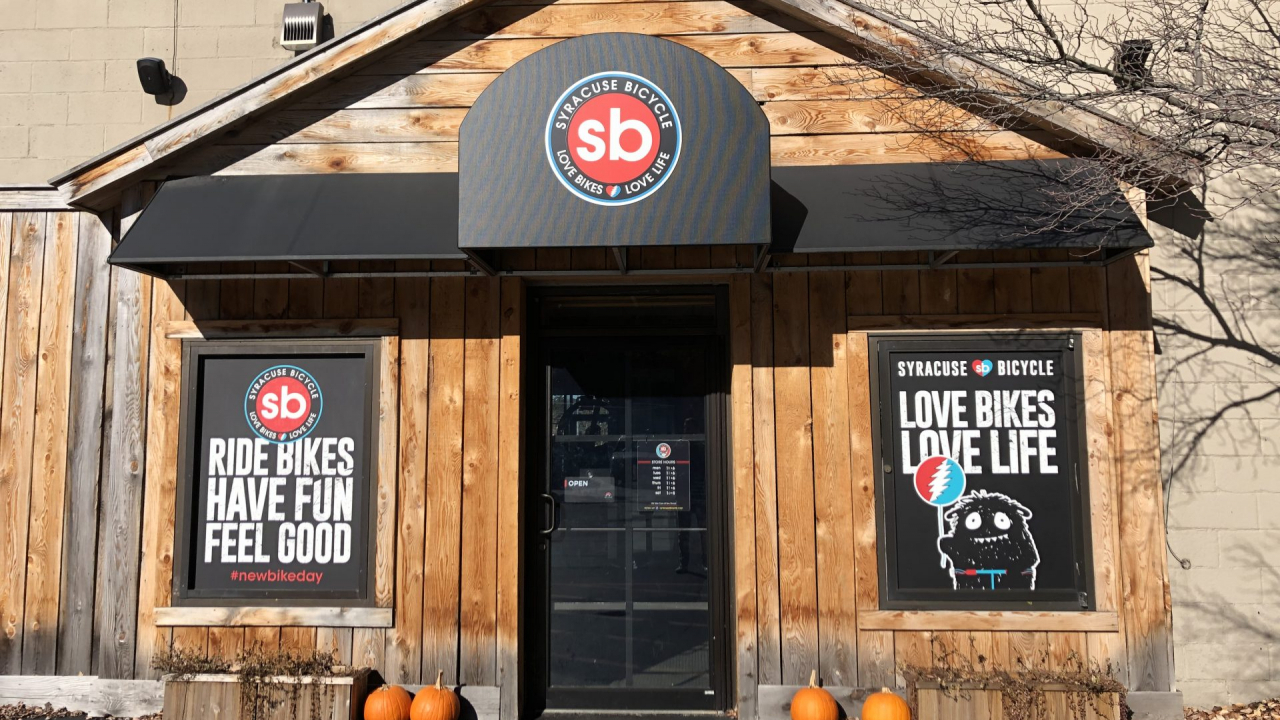 Pictured is the exterior of the entrance of the store. The entrance consists of vertical wooden planks with the shops logo at the top of the wall with two other signs on each side of the front door. The sign on the left reads “Ride Bikes, Have Fun, Feel Good”. The sign on the right reads “Love Bikes, Love Life”.