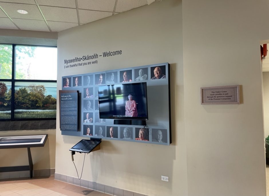 A television screen playing a welcome video is surrounded by photos of local Indigenous people. The welcoming video that greets frequenters of the Skä•noñh Great Law of Peace Center upon arrival. It begins with the phrase "Nyawenha Skanonh" or "I am thankful that you are well."