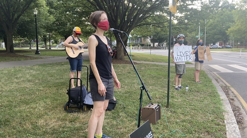 Women singing at microphone while another woman plays guitar. The singer's husband and daughter hold signs nearby.