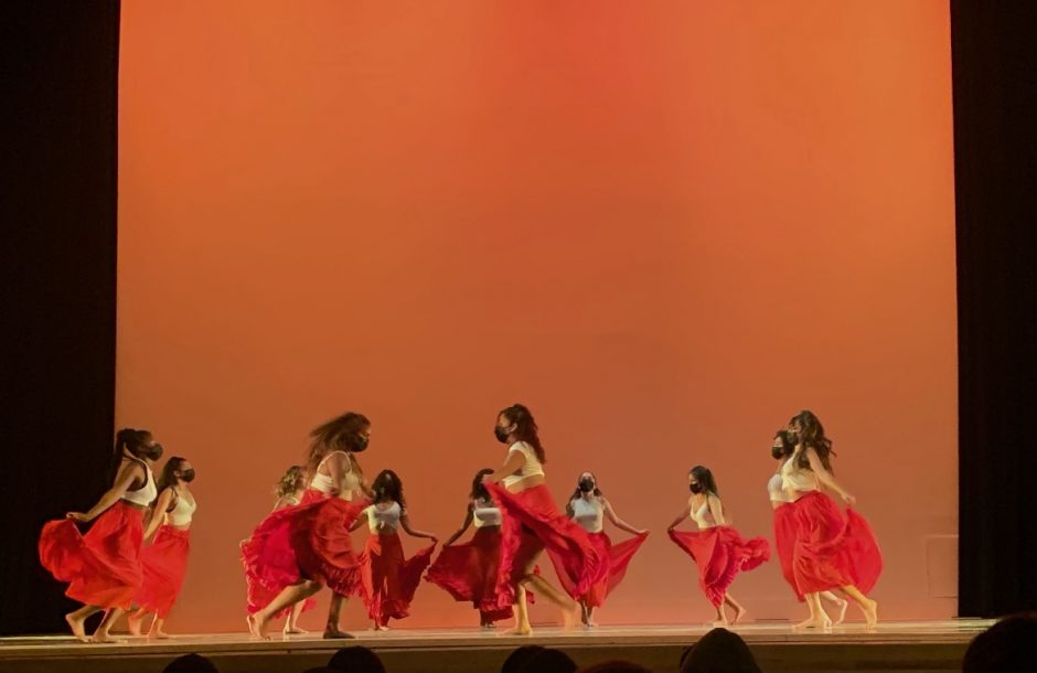 11 young women in long red skirts dancing on a stage.