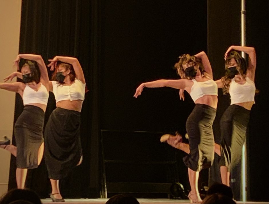 Four Raices women swing a leg and arm up midst dance. They are wearing white tops and flowy black skirts.