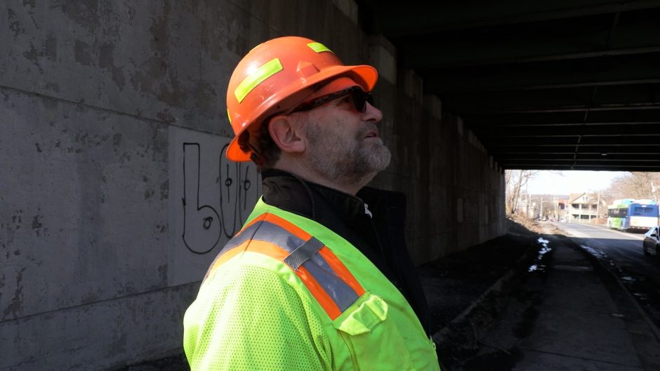 A man in safety gear looks up and to the right underneath an overpass.