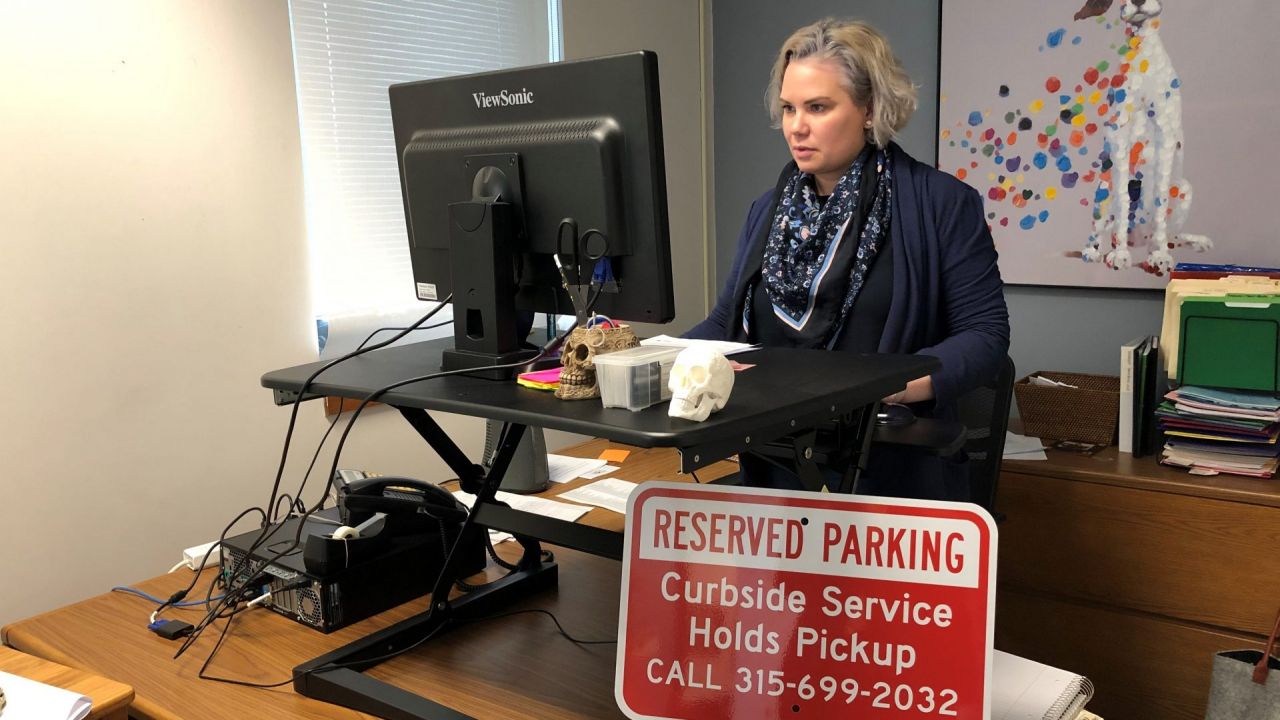A woman uses a computer at a standing desk. A red sign that reads "Reserved Parking: Curbside Service Holds Pickup" is placed in front of her computer.