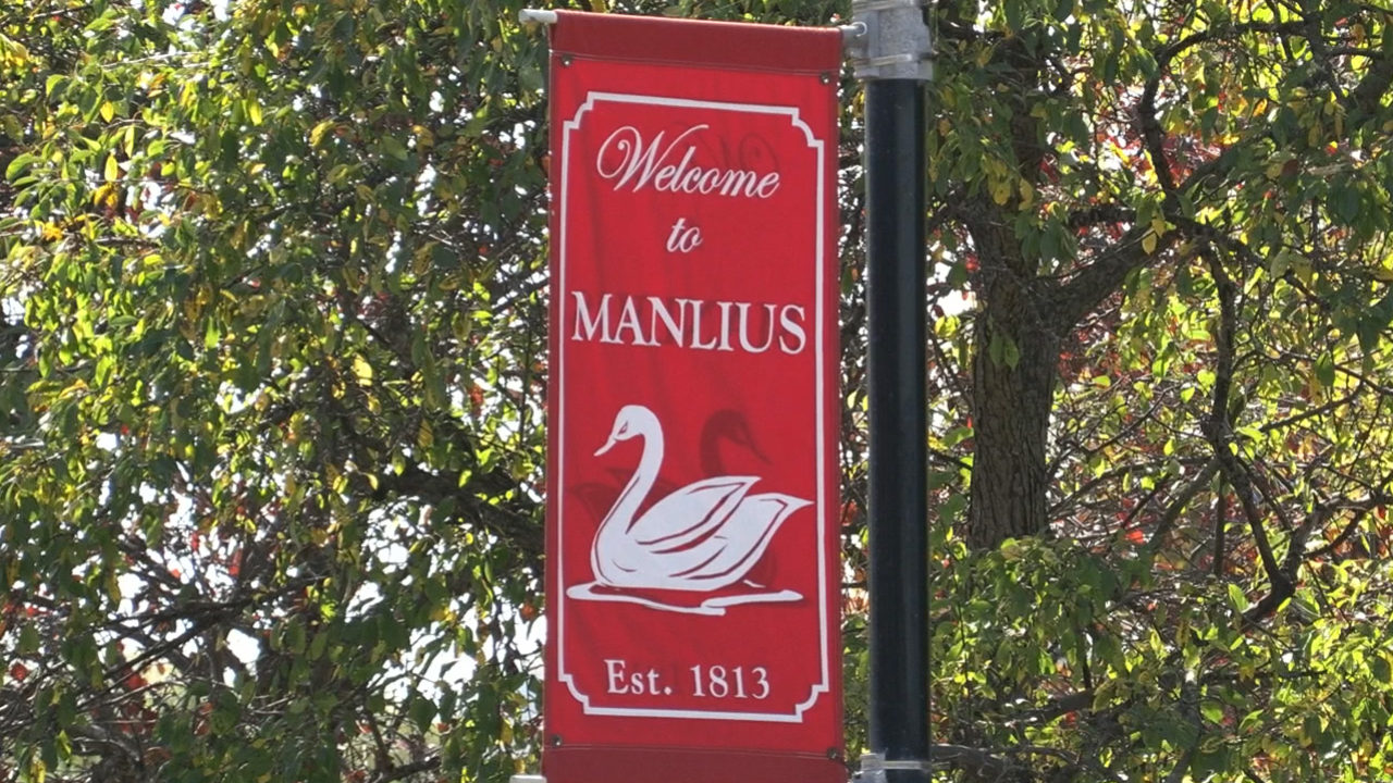 A red sign with a swan on it that reads "Village of Manlius." There is a tree behind the sign.