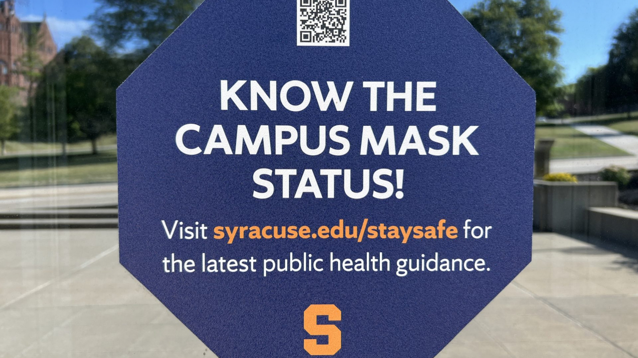 A sign displays the information for the mask status outside a Syracuse University building.