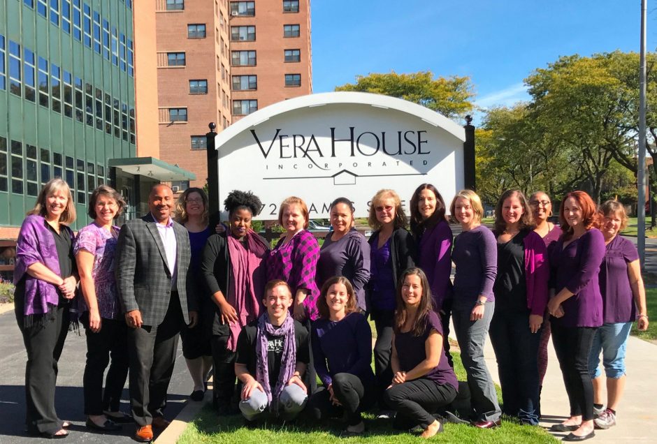 Last year's group photo of Vera House staff, dressed in purple and posing in front of the Vera House sign.