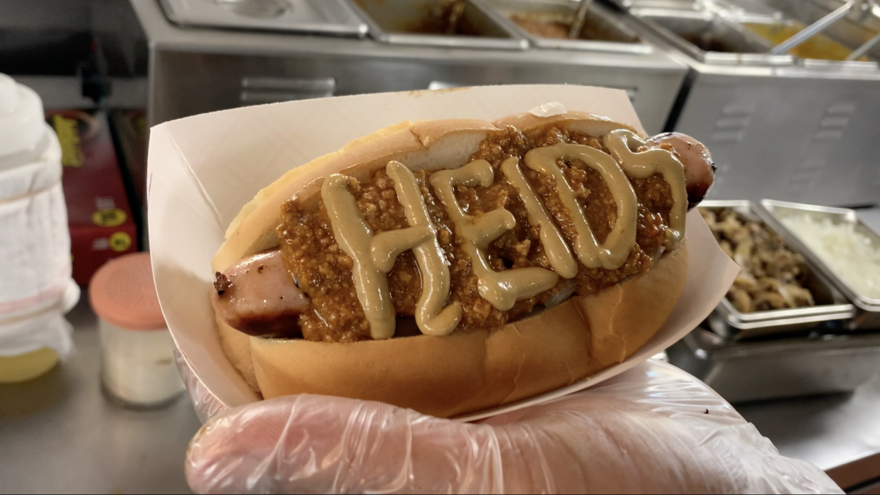 A hot dog with onions, chili and spells out "HEIDS" with honey mustard.