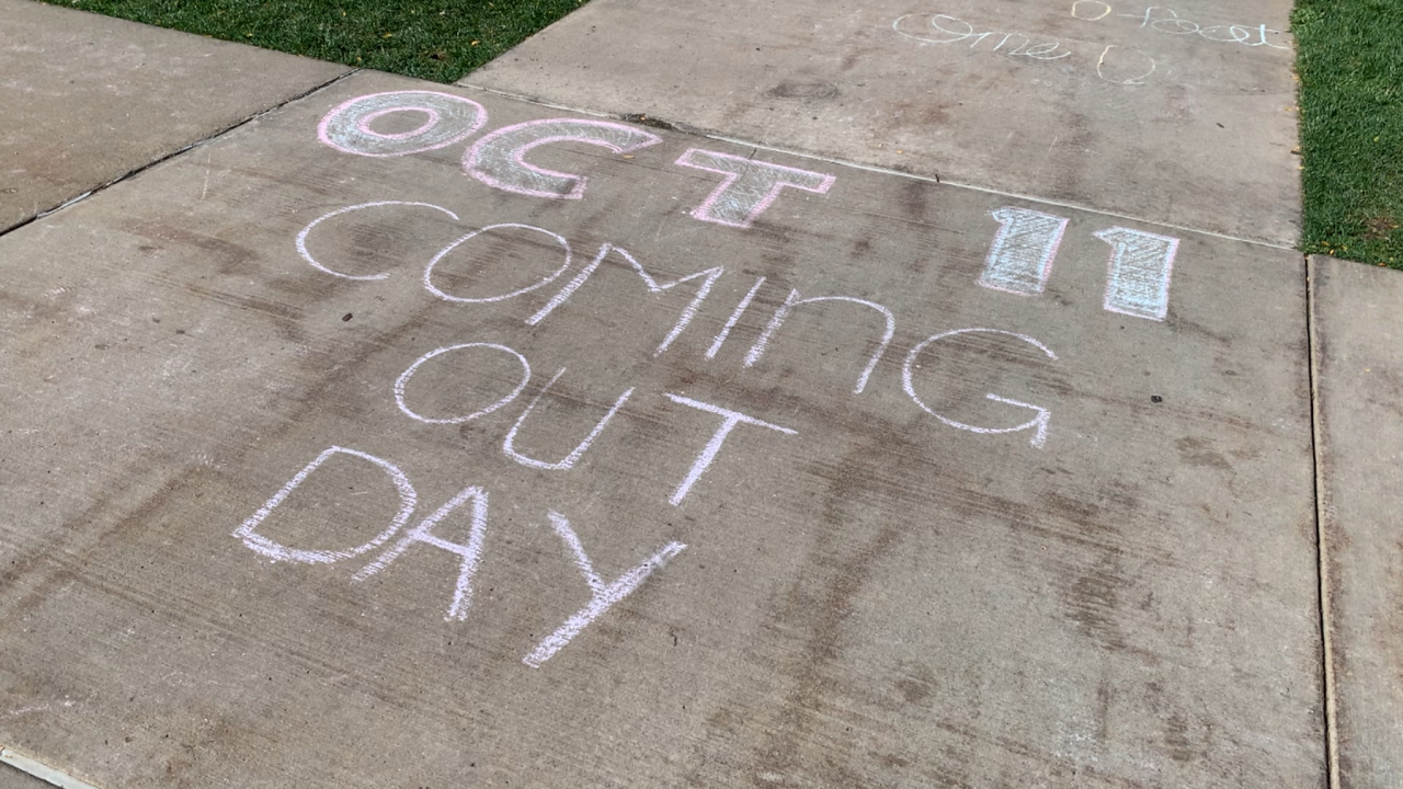 A chalk drawing on the sidewalk that says "Oct 11 Coming Out Day"