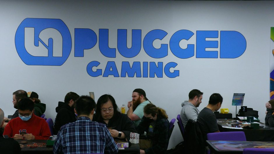 A large sign of Unplugged Gaming in blue lettering with people sitting at tables underneath.