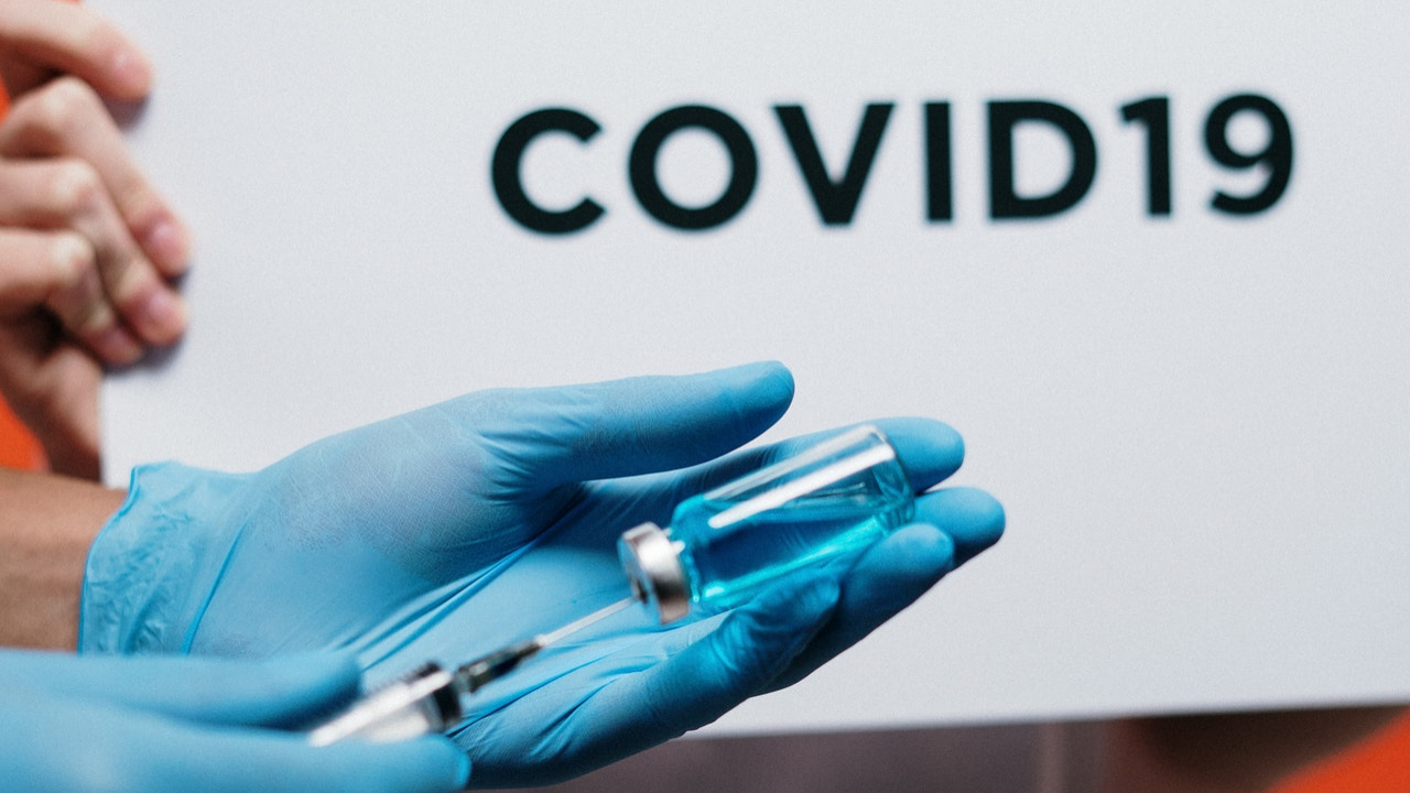 A syringe containing a vaccine is held in front of a piece of paper that reads, "COVID-19."