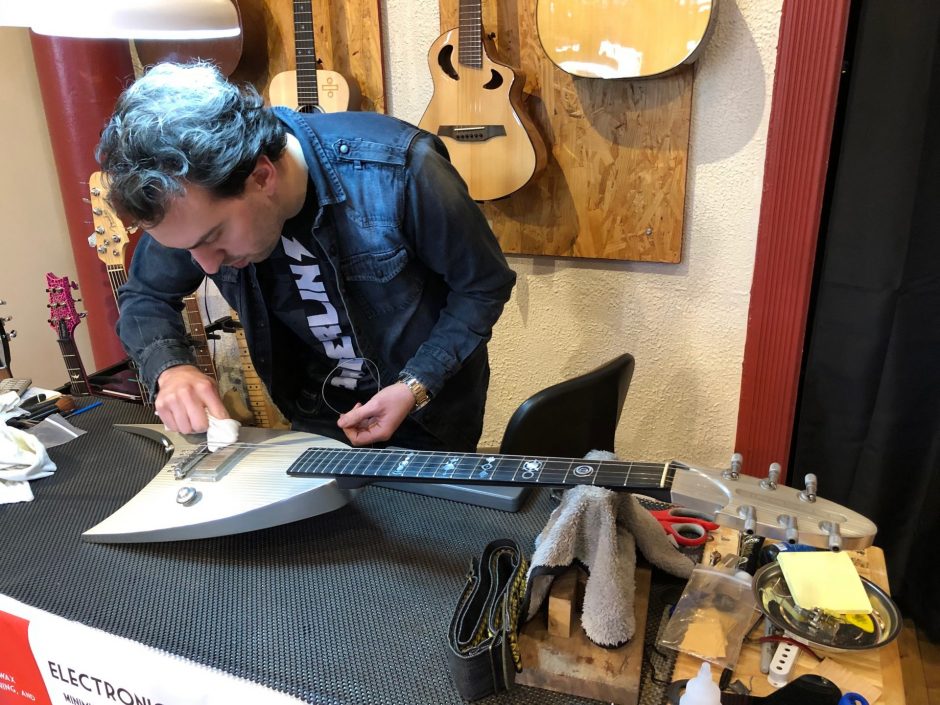 A man in a denim jacket polishes a silver V-shaped guitar on a table surrounded by other guitars.
