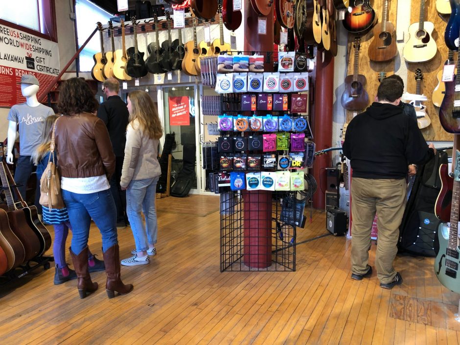 Three men, a woman, and two girls look at acoustic and electric guitars displayed on the walls and the floors of a wood-paneled store; a display showing guitar strings occupies the center of the floor.