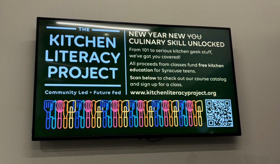 A TV in Salt City Market is showing a graphic advertising the Kitchen Literacy Project
